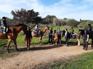 Um grupo de pessoas monta cavalos e pôneis ao longo de um caminho de terra em uma área gramada com árvores ao fundo. Algumas pessoas usam capacetes e o tempo parece ensolarado com céu azul.